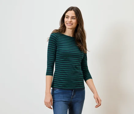 Une femme aux longs cheveux bruns porte un T-shirt à manches longues rayé vert et noir et un jean sur un fond blanc.