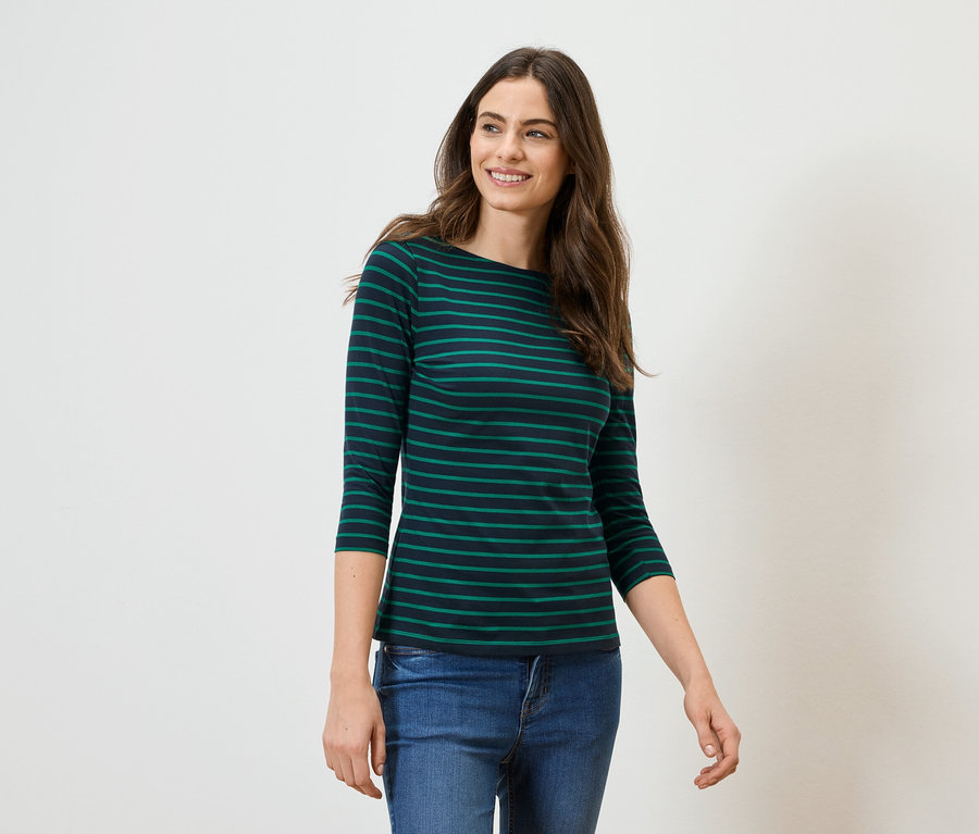 Une femme aux longs cheveux bruns porte un T-shirt à manches longues rayé vert et noir et un jean sur un fond blanc.