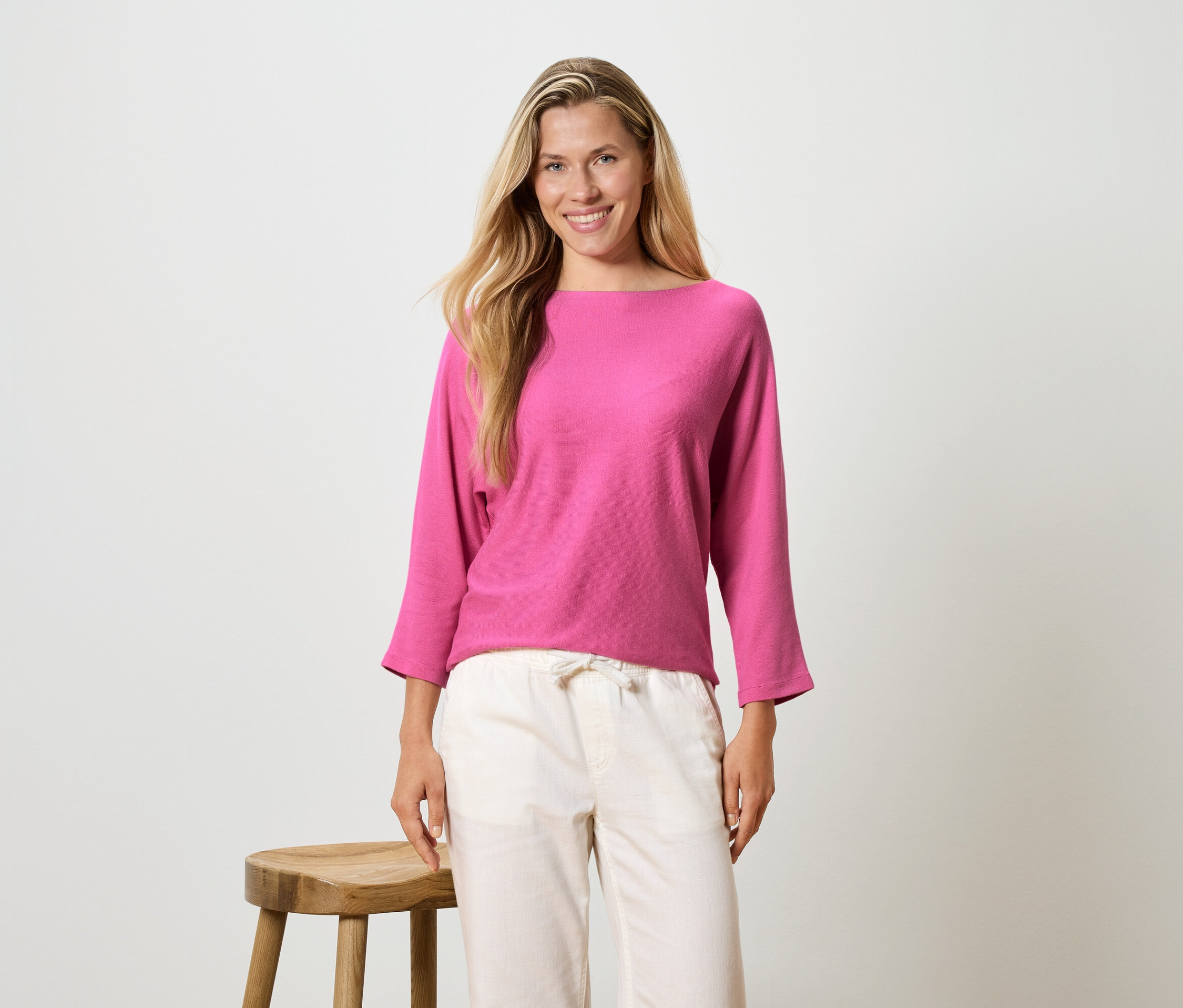 Une femme aux longs cheveux blonds porte un haut à manches longues rose et un pantalon clair, et se tient à côté d'un tabouret.