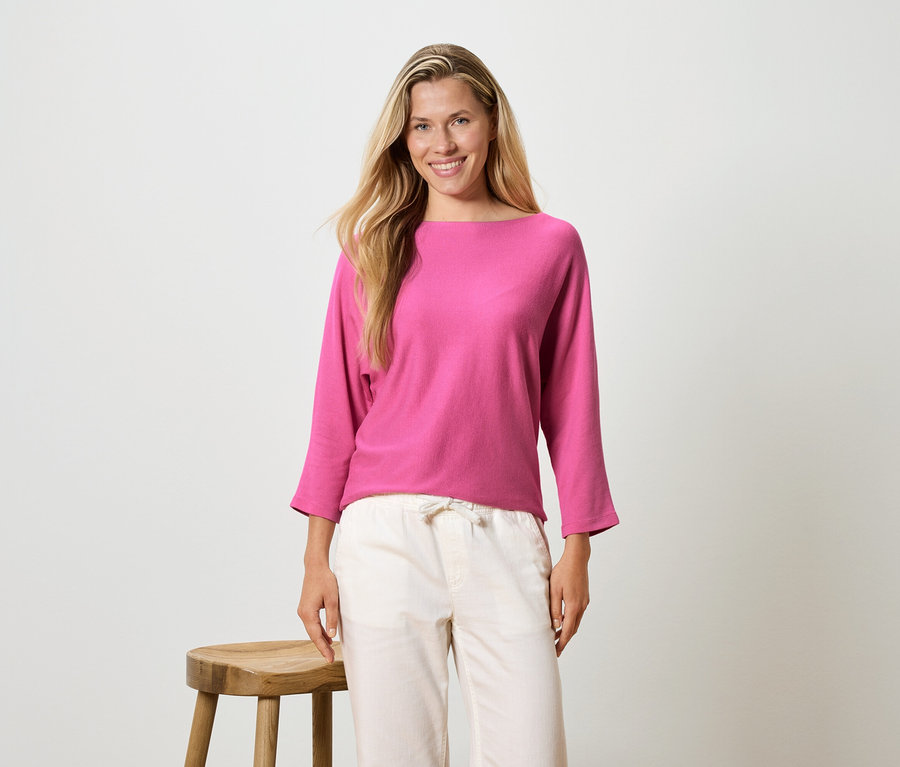 Une femme aux longs cheveux blonds porte un haut à manches longues rose et un pantalon clair, et se tient à côté d'un tabouret.