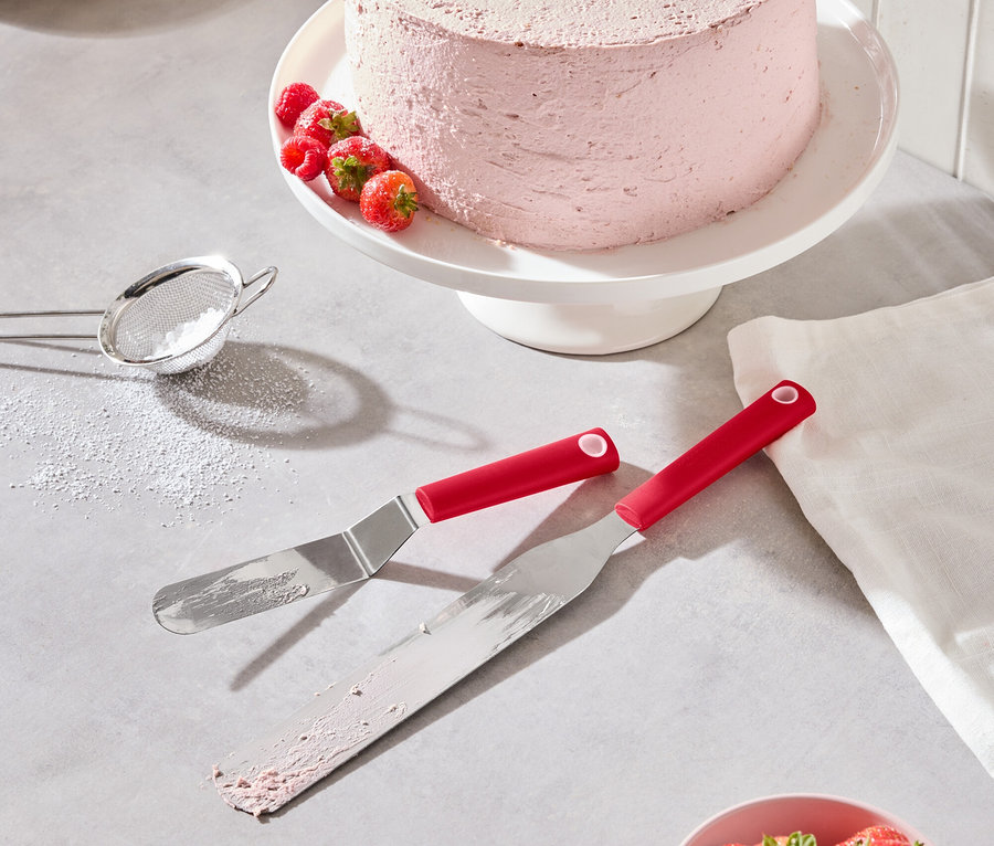 Un gâteau rose aux fraises et framboises est posé sur un présentoir à gâteau blanc, avec deux spatules rouges, un bol de fraises et un tamis à sucre glace.