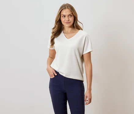 Une femme aux longs cheveux ondulés porte un t-shirt ample crème à manches courtes et col en V, ainsi qu'un pantalon bleu marine, avec une main dans la poche.