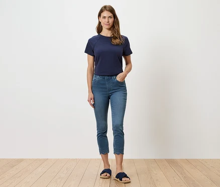 Femme portant un t-shirt bleu marine et un jean bleu avec broderie ajourée à l'ourlet, posant devant un mur blanc.