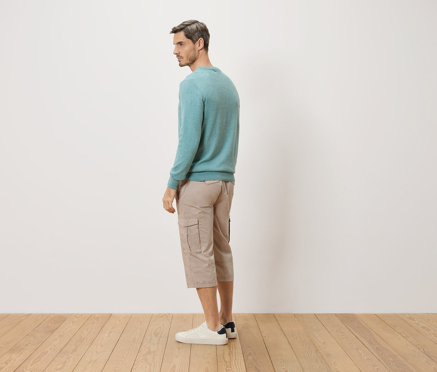 Un homme se tient de côté, portant un pull turquoise, un short beige et des chaussures blanches.