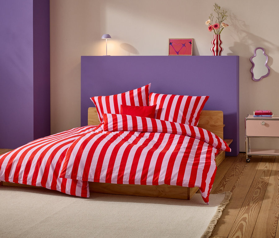 Lit avec des draps rayés rouge et blanc et plusieurs oreillers devant un mur violet.
