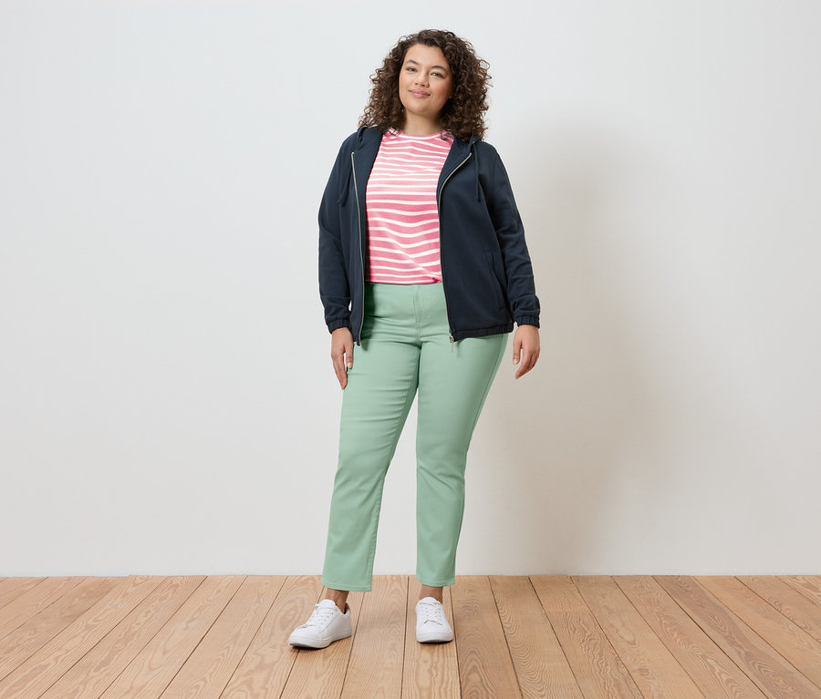 Une femme se tient dans un environnement lumineux, portant une veste bleu marine, un haut rayé, un pantalon vert clair et des baskets blanches.