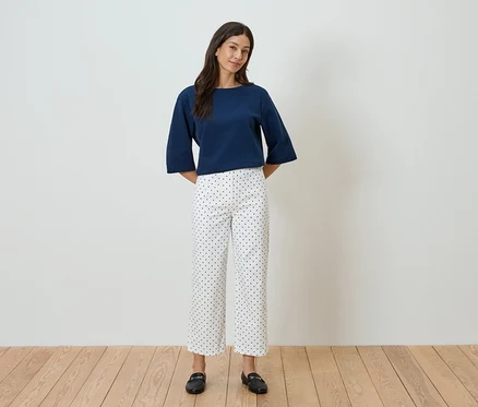 Une femme se tient devant un mur blanc, portant un haut bleu et un pantalon blanc à pois noirs.