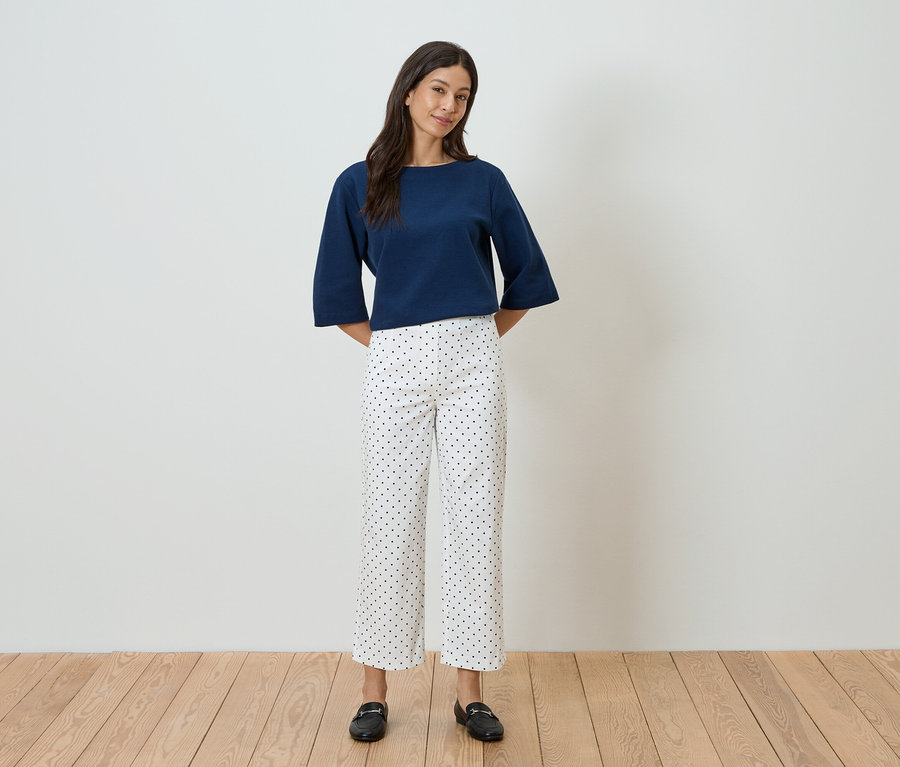 Une femme se tient devant un mur blanc, portant un haut bleu et un pantalon blanc à pois noirs.