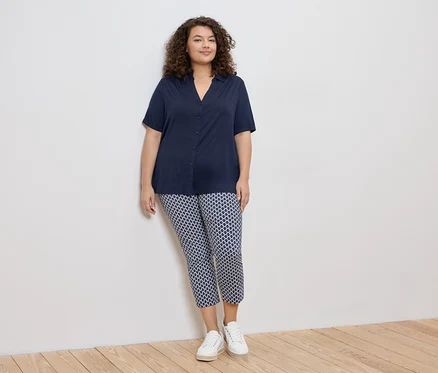 Femme aux cheveux bouclés portant un chemisier bleu marine et un pantalon à motifs avec des baskets blanches, appuyée contre un mur blanc.