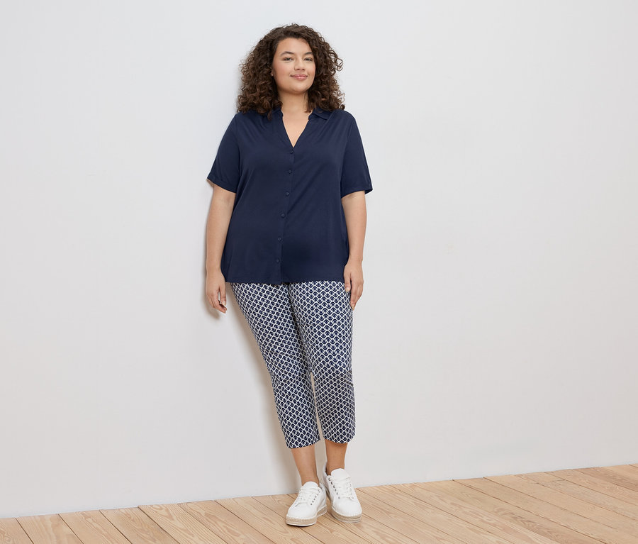Femme aux cheveux bouclés portant un chemisier bleu marine et un pantalon à motifs avec des baskets blanches, appuyée contre un mur blanc.