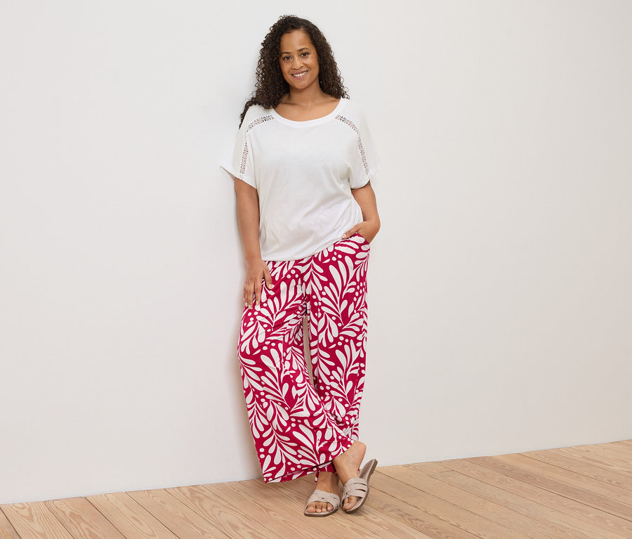 Une femme aux cheveux bouclés, portant un t-shirt blanc et un pantalon rouge et blanc, est appuyée contre un mur.