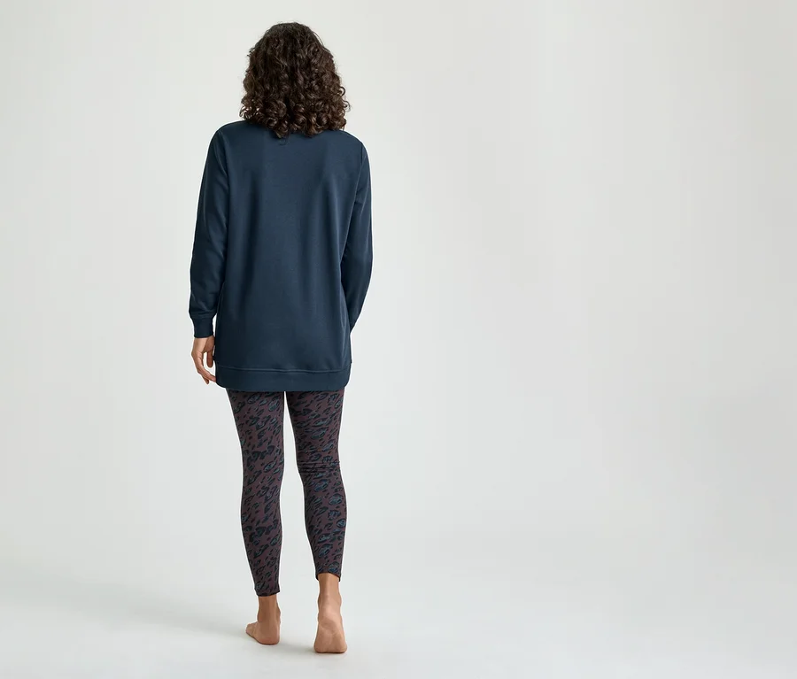Une femme vue de dos, portant un haut à manches longues bleu foncé et un legging foncé à motifs, debout pieds nus.