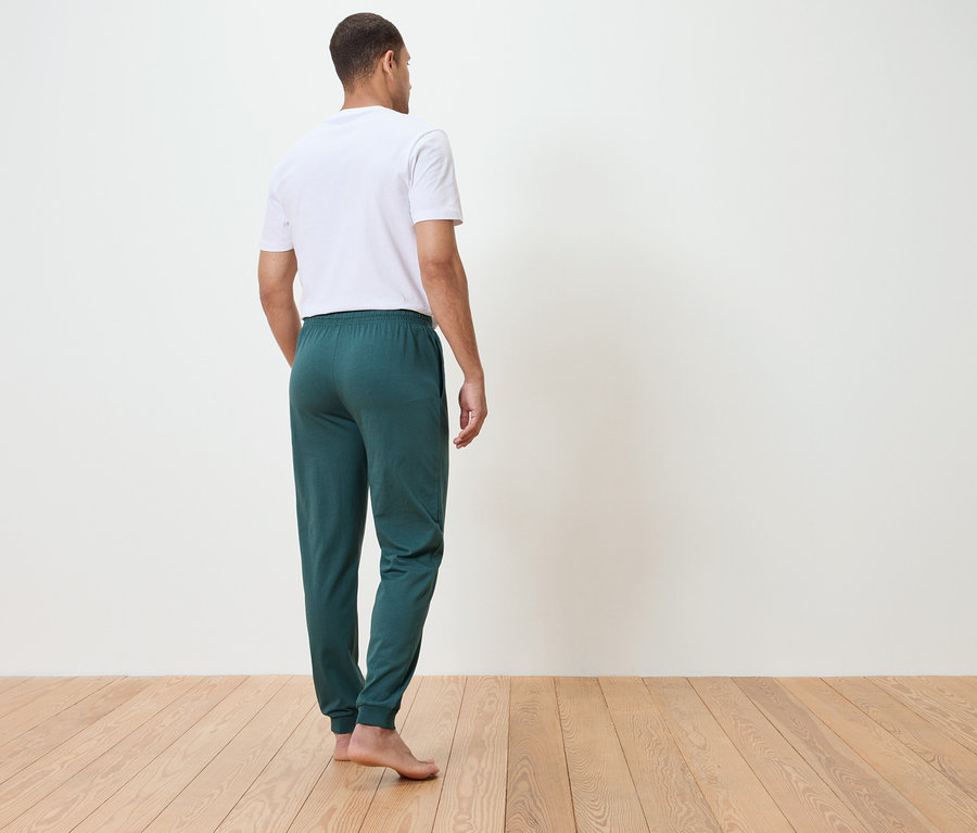 Homme vu de dos portant un t-shirt blanc et un pantalon de jogging vert, debout sur un plancher en bois devant un mur blanc.