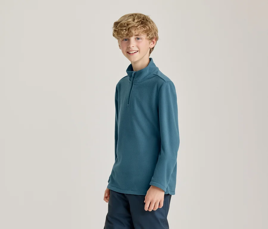 Un jeune garçon souriant aux cheveux blonds bouclés porte un haut à manches longues sarcelle avec un col à demi-zip et un pantalon bleu foncé, debout devant un fond clair.