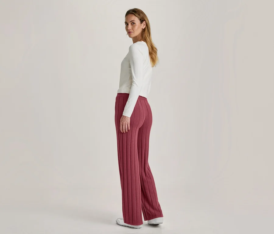 Une femme se tient dos à la caméra, regardant par-dessus son épaule. Elle porte un haut blanc à manches longues, un pantalon côtelé, large, couleur baie et des chaussures blanches.