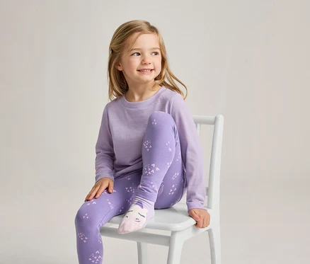 Une jeune fille aux cheveux blonds est assise sur une chaise blanche, souriant et regardant vers la droite. Elle porte un haut à manches longues violet clair, un legging violet avec un motif violet plus clair et des chaussettes rose clair avec un motif de visage.