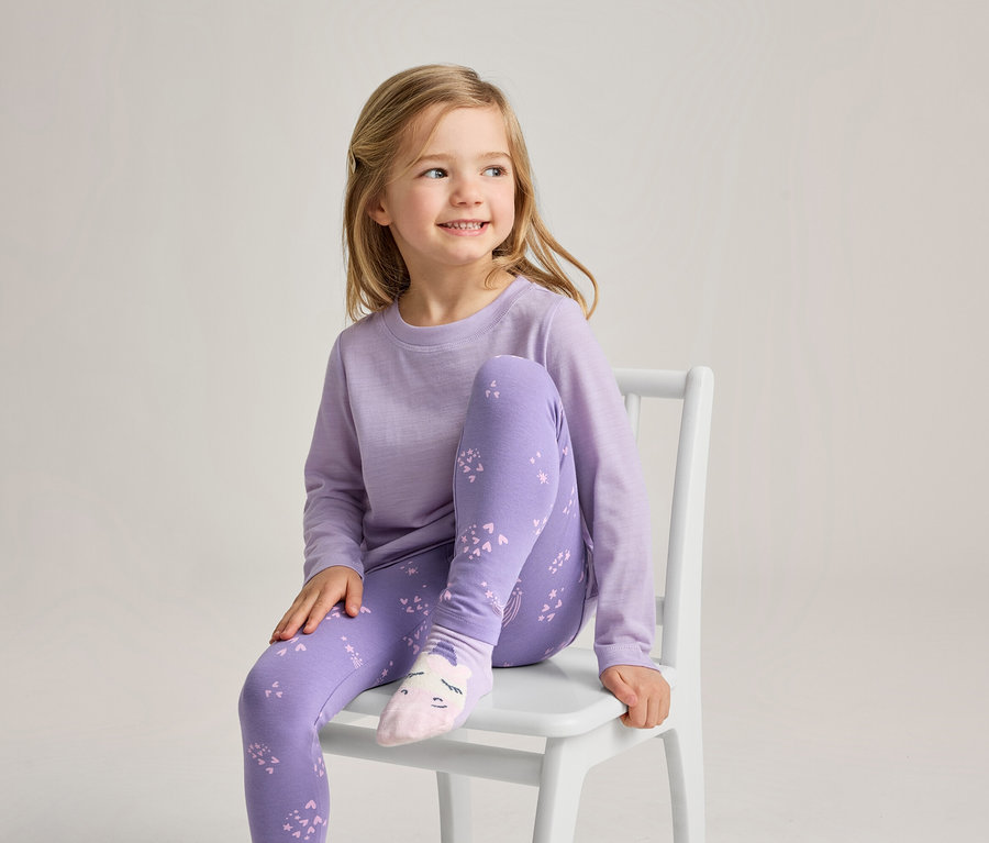 Une jeune fille aux cheveux blonds est assise sur une chaise blanche, souriant et regardant vers la droite. Elle porte un haut à manches longues violet clair, un legging violet avec un motif violet plus clair et des chaussettes rose clair avec un motif de visage.