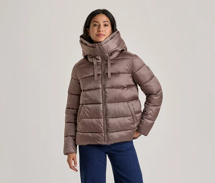 Femme portant une doudoune marron avec un col montant et une capuche, et un jean bleu, debout les mains dans les poches.