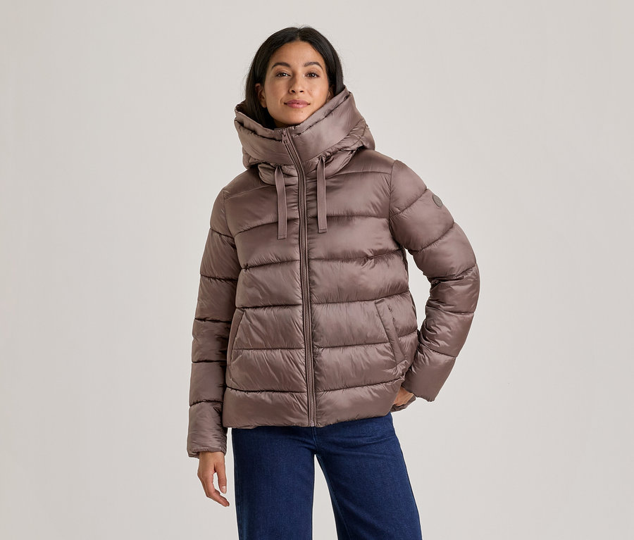 Femme portant une doudoune marron avec un col montant et une capuche, et un jean bleu, debout les mains dans les poches.