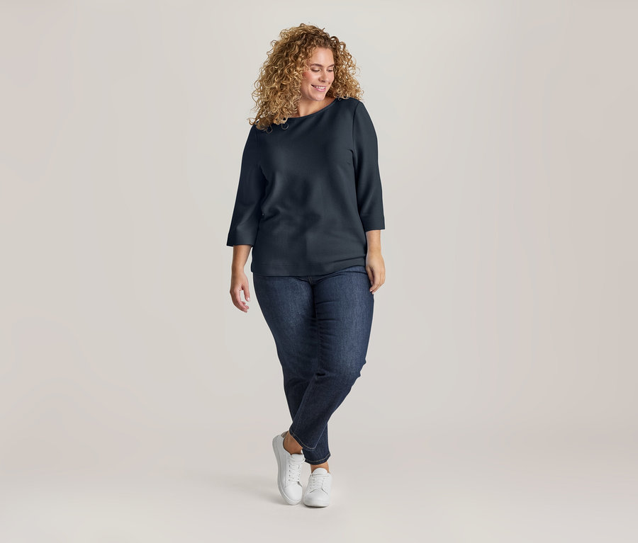 Femme souriante aux cheveux bouclés, portant un haut bleu marine à col bateau, un jean foncé et des baskets blanches, debout les jambes croisées.