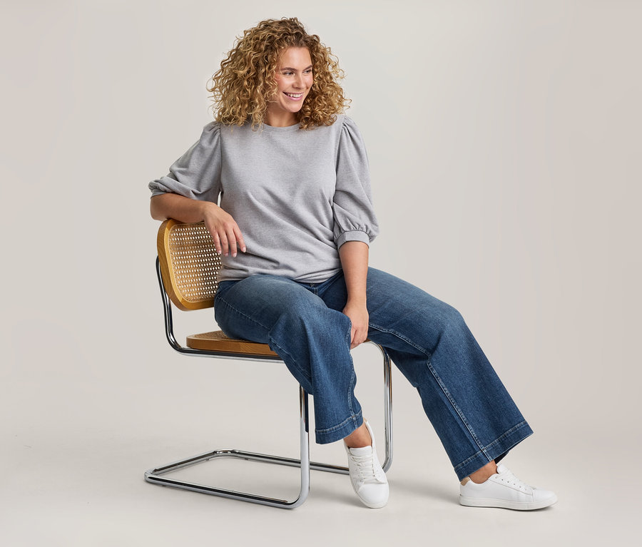 Femme aux cheveux blonds bouclés assise sur une chaise moderne avec un dossier en cannage et des pieds chromés. Elle porte un sweat-shirt gris à manches bouffantes, un jean bleu large et des baskets blanches. Elle sourit et regarde vers la droite, son bras gauche reposant sur le dossier de la chaise.