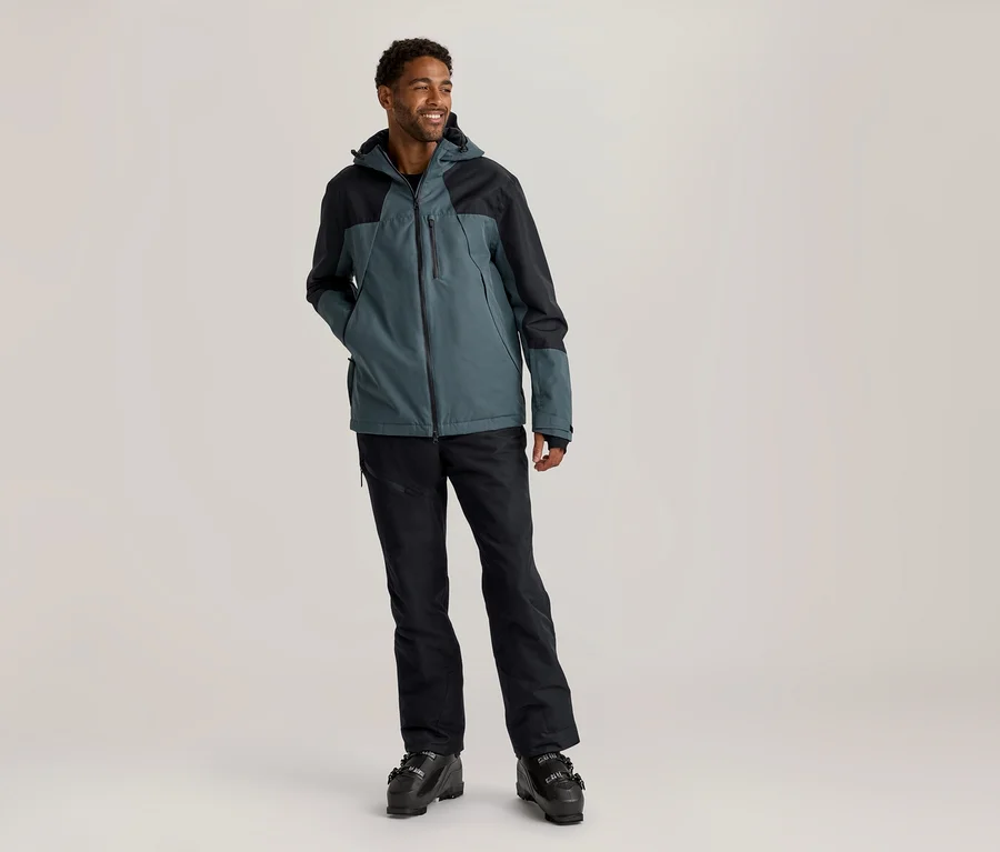 Homme debout, souriant et regardant vers la droite, portant une veste de ski turquoise et noire, un pantalon de ski noir et des chaussures de ski noires.