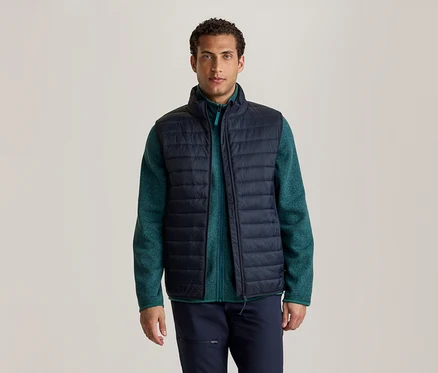 Un homme se tient de face, portant une doudoune sans manches bleu foncé sur une veste polaire verte et un pantalon bleu foncé.