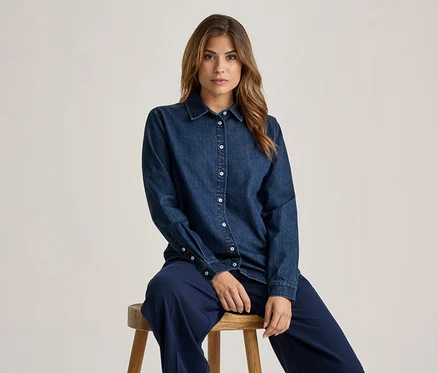 Femme aux longs cheveux bruns assise sur un tabouret en bois, portant une chemise en jean bleu foncé avec des boutons blancs et un pantalon bleu foncé, regardant devant elle.