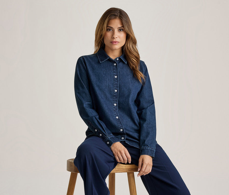 Femme aux longs cheveux bruns assise sur un tabouret en bois, portant une chemise en jean bleu foncé avec des boutons blancs et un pantalon bleu foncé, regardant devant elle.