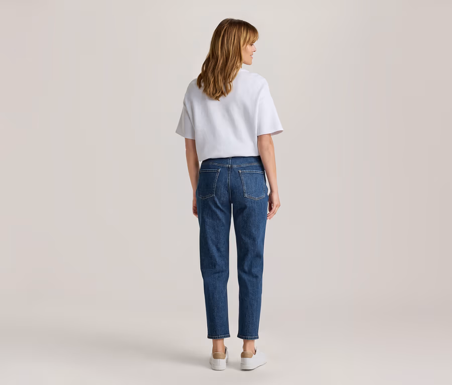 Femme de dos, vêtue d'un t-shirt blanc, d'un jean bleu et de baskets blanches, regardant par-dessus son épaule droite.