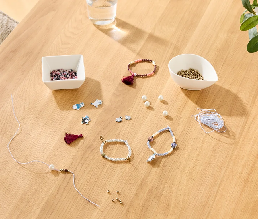 Matériel de fabrication de bracelets, comprenant des perles dans des bols, des perles lâches, des breloques d'animaux, des pompons, du fil et des bracelets finis/partiellement faits sur une table en bois.