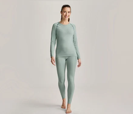 Femme souriante en haut à manches longues vert clair et legging assorti avec coutures foncées, pieds nus.