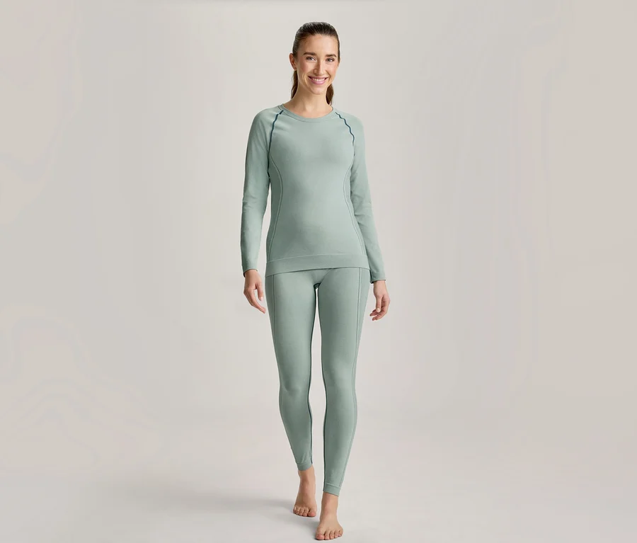 Femme souriante en haut à manches longues vert clair et legging assorti avec coutures foncées, pieds nus.