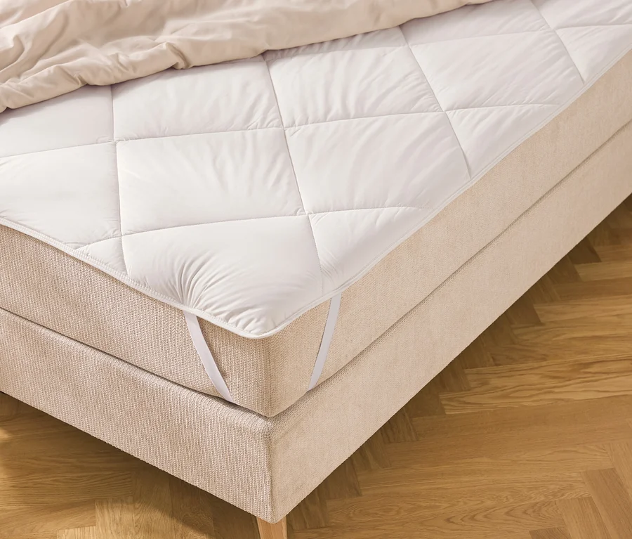 Un lit avec un cadre en tissu beige et un surmatelas blanc matelassé, fixé par des sangles élastiques. Une couette claire est pliée en haut, le lit repose sur un parquet à chevrons en bois.