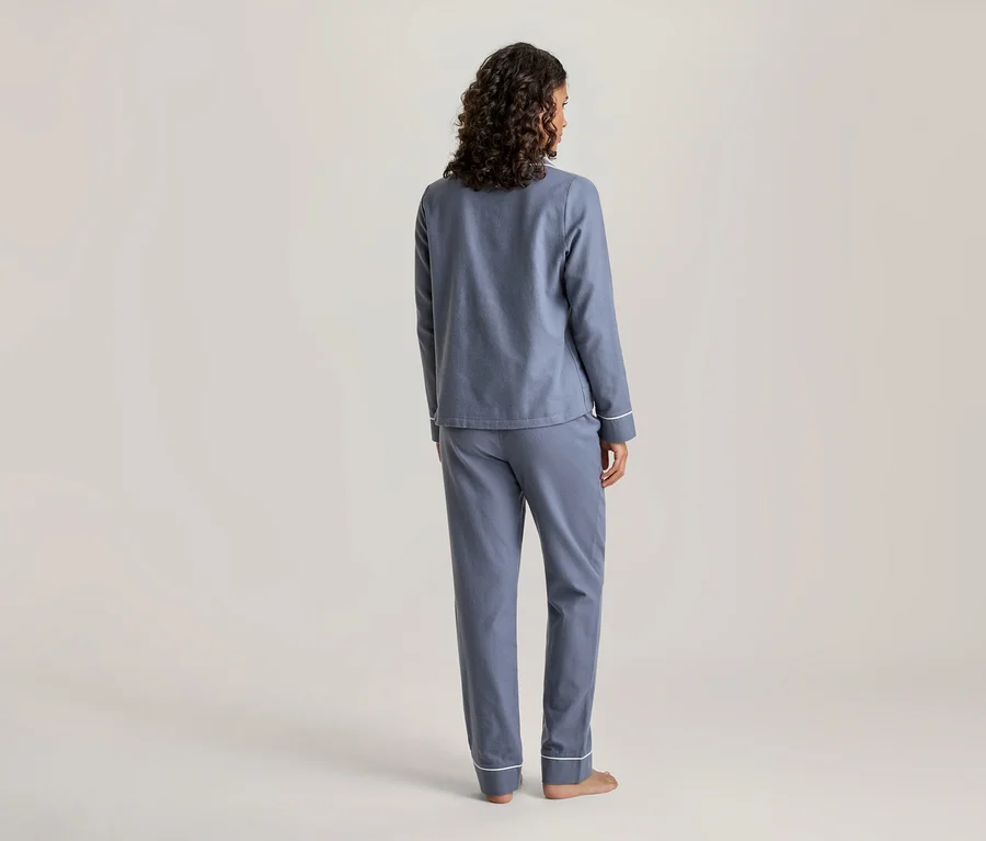 Une personne aux cheveux bruns bouclés, vue de dos, porte un ensemble de pyjama bleu avec des passepoils blancs aux poignets et aux ourlets du pantalon. La personne est pieds nus et se tient devant un fond clair uni.