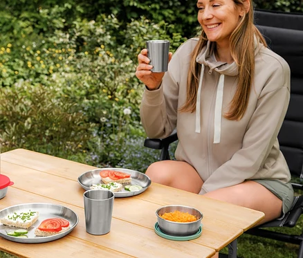 Une femme souriante en veste en sweat beige et short fonctionnel de plein air est assise à une table de camping, tenant un gobelet de camping. Sur la table, deux assiettes de camping avec des sandwichs et un autre gobelet de camping.