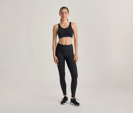 Une femme se tient debout, vêtue d'un soutien-gorge de sport noir, d'un legging noir et de chaussures de sport noires, sur un fond clair.