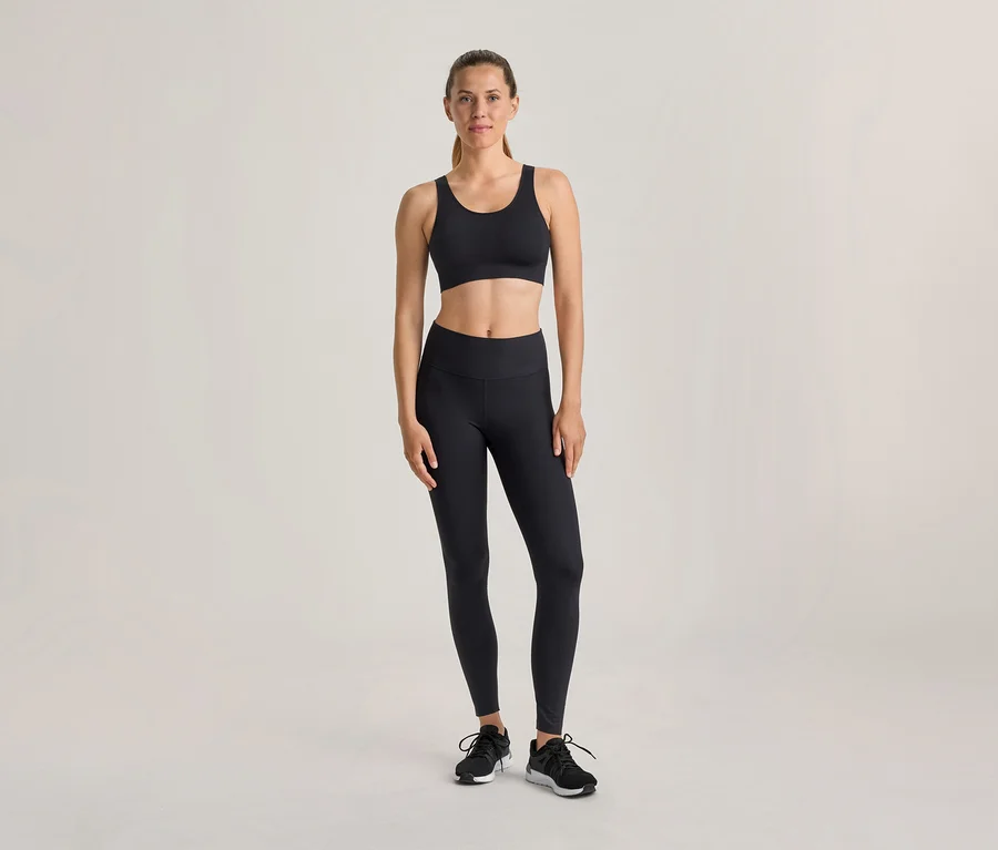Une femme se tient debout, vêtue d'un soutien-gorge de sport noir, d'un legging noir et de chaussures de sport noires, sur un fond clair.