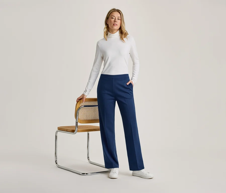 Une femme vêtue d'un col roulé blanc à manches longues, d'un pantalon large bleu marine et de baskets blanches se tient à côté d'une chaise en bois à cadre chromé, la main posée sur son dossier.