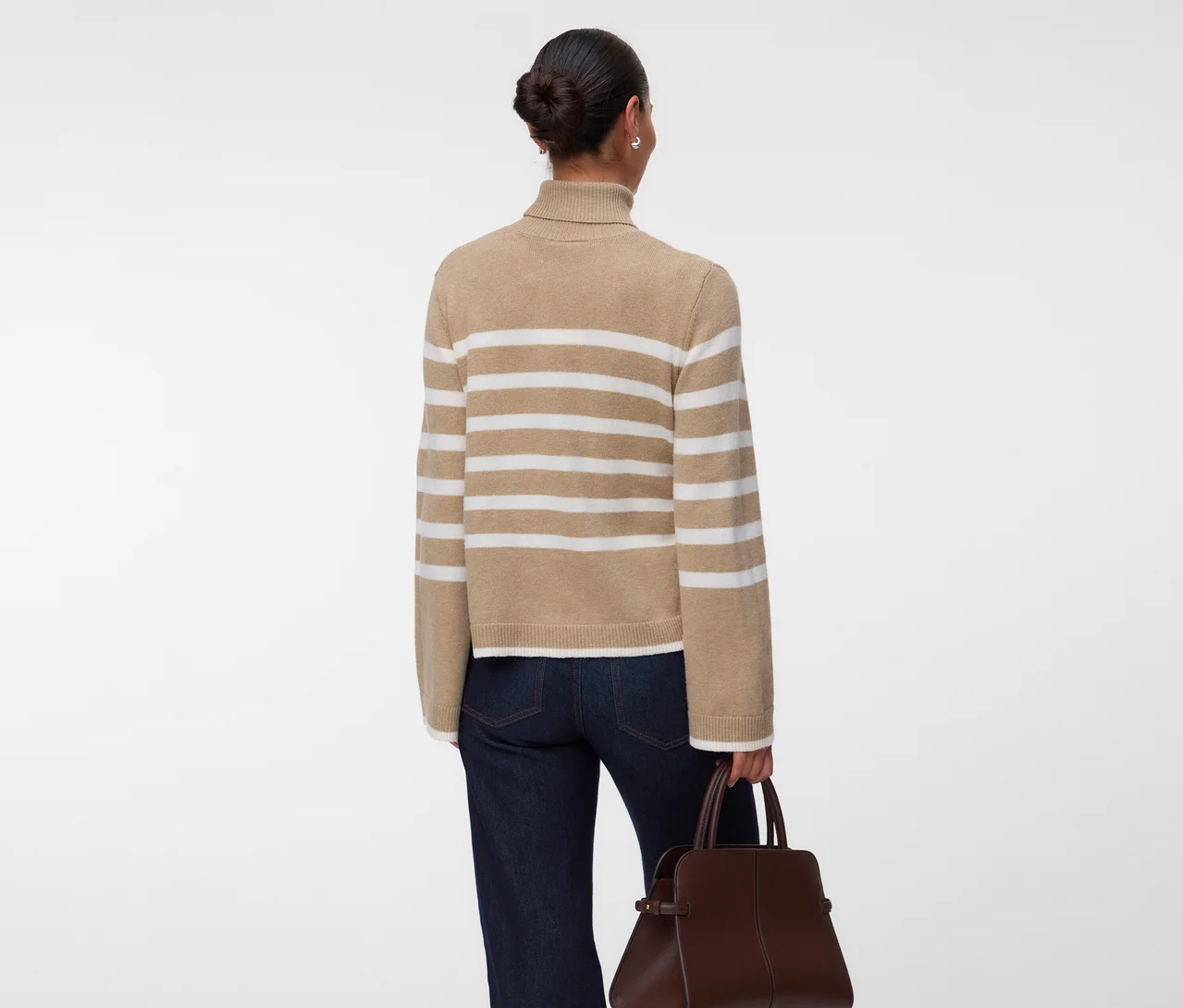 Personne de dos portant un pull à col roulé beige à rayures blanches, un jean foncé et tenant un sac à main marron.