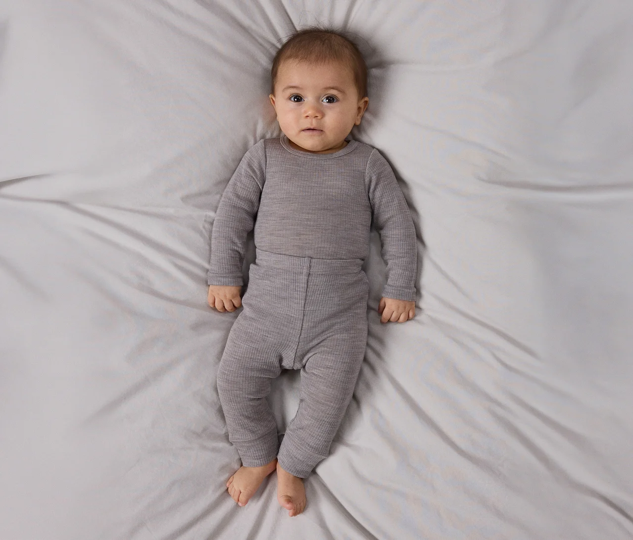Bébé allongé sur le dos sur un drap gris clair, portant un haut côtelé gris à manches longues et un pantalon assorti, regardant la caméra.