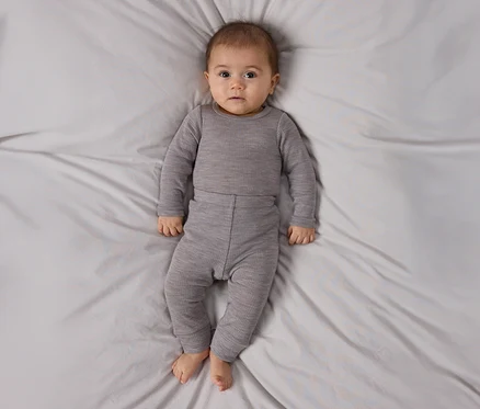 Bébé allongé sur le dos sur un drap gris clair, portant un haut côtelé gris à manches longues et un pantalon assorti, regardant la caméra.