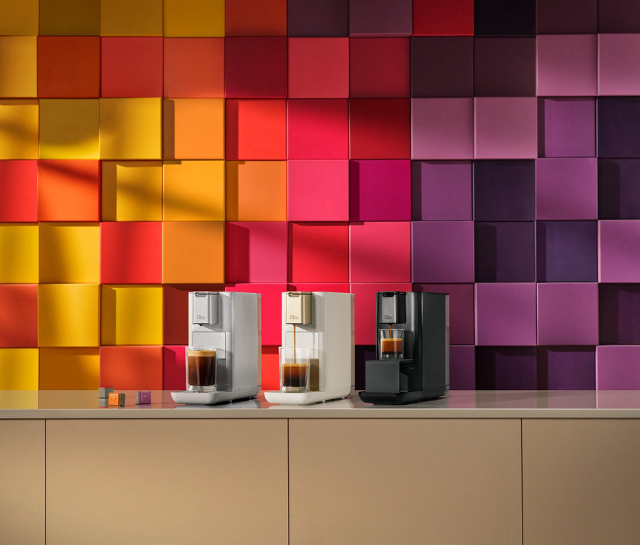 Trois machines à café, gris clair, crème et noir, sont posées sur un comptoir beige. La machine crème verse du café dans un verre. Derrière elles, un mur de carreaux carrés présente un dégradé de couleurs allant du jaune et orange à gauche au rouge, rose et violet à droite. Des capsules de café sont également sur le comptoir.