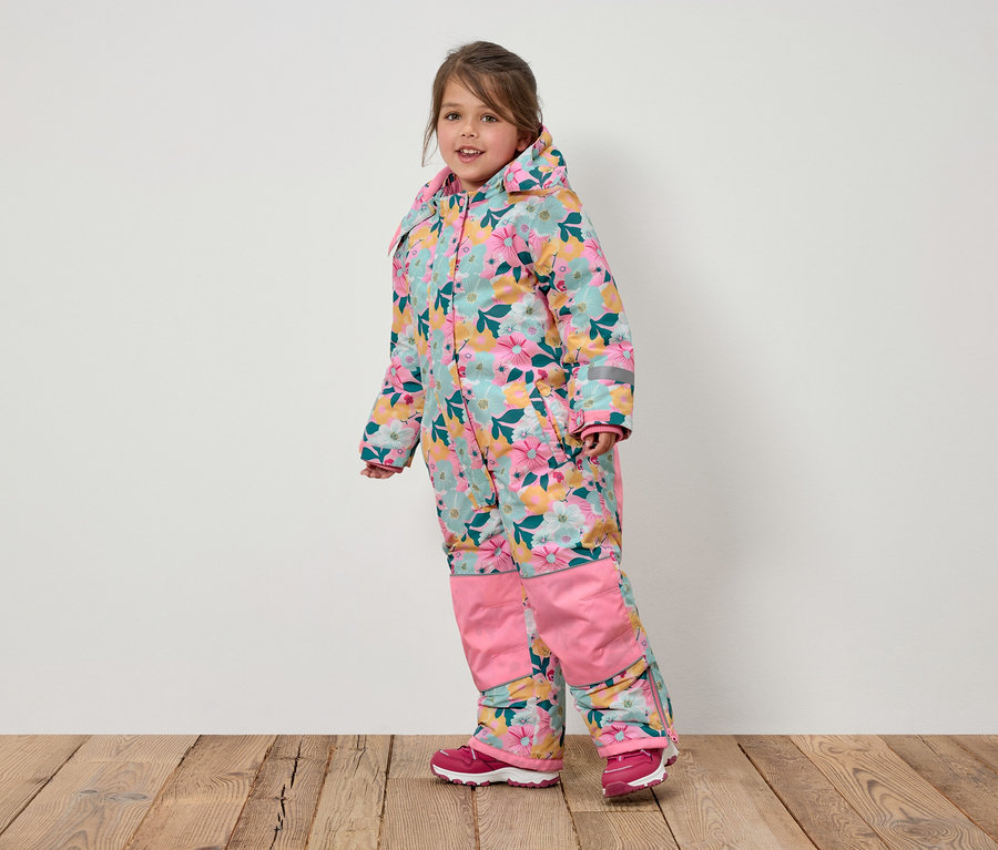 Une fillette souriante vêtue d'une combinaison à fleurs avec capuche et des chaussures roses se tient sur un plancher en bois.