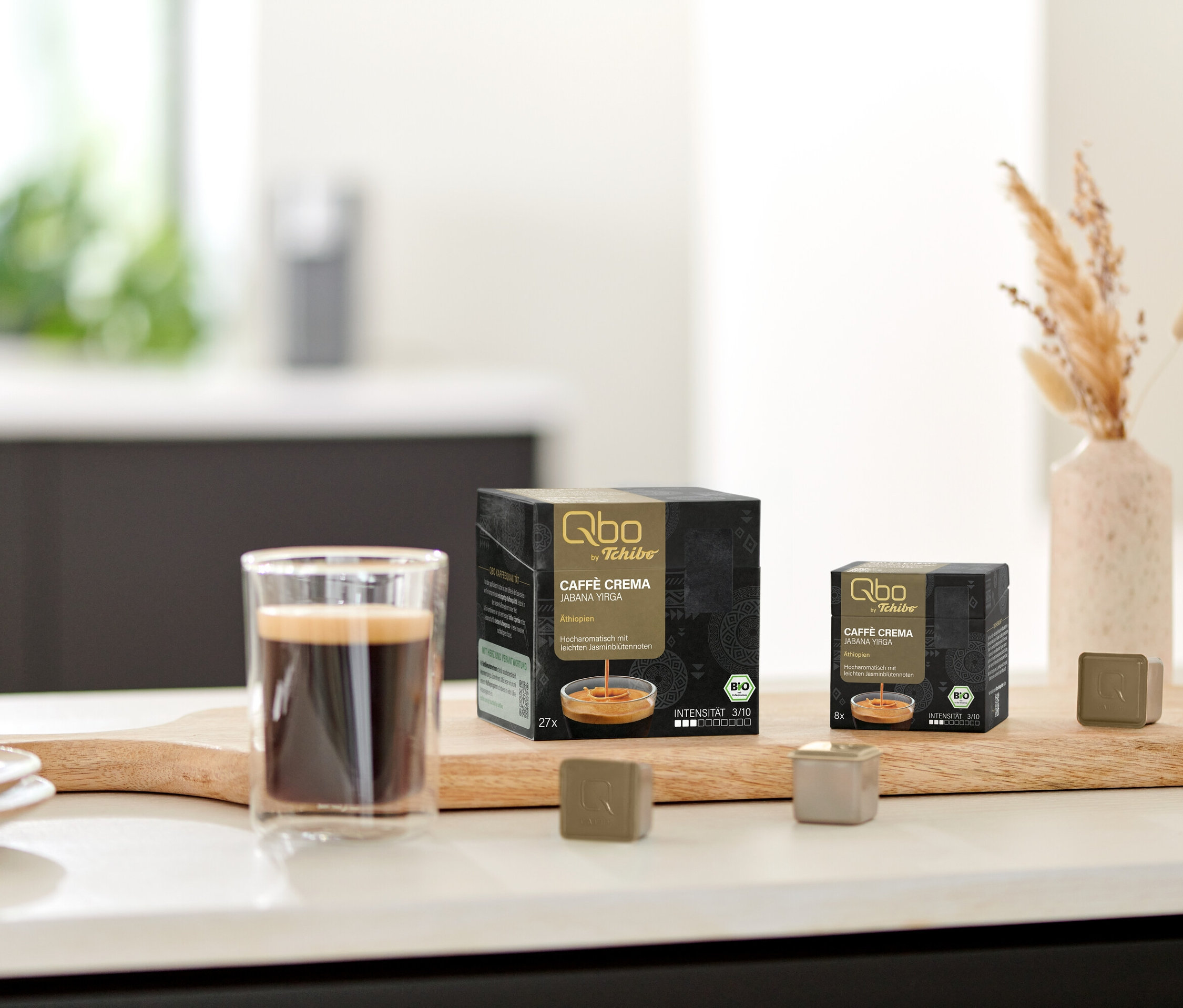 Sur une planche se trouvent une tasse de café, un paquet de Qbo Caffè Jabana Yirga, 27 capsules et un paquet de Qbo Caffè JABANA YIRGA - 8 Kapseln.