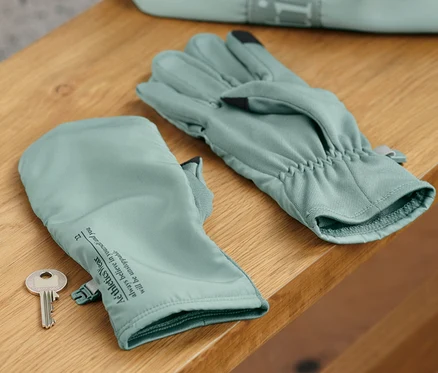 Sur une étagère en bois reposent des gants de course à pied avec rabats et une clé, avec un sac en arrière-plan.
