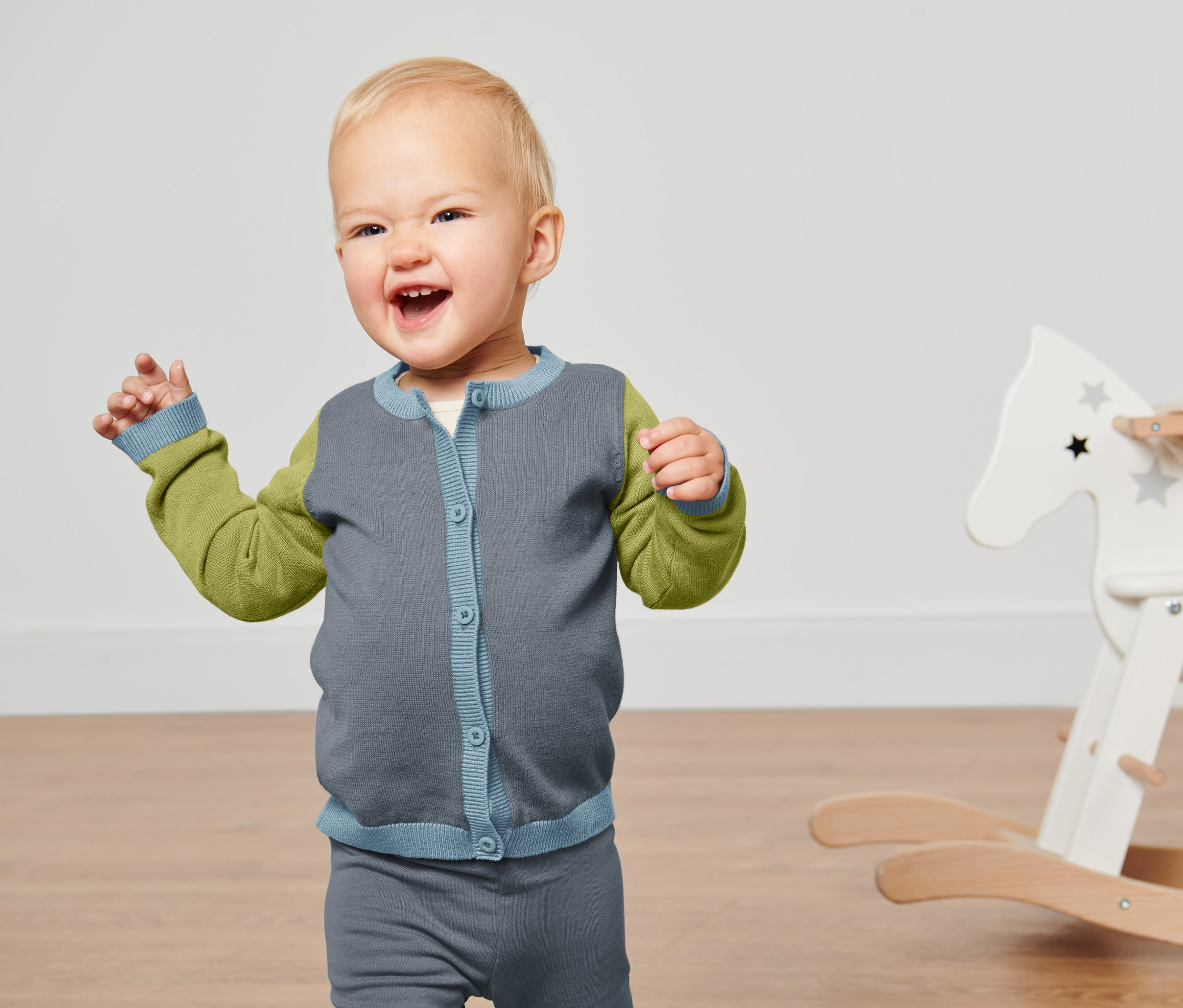 Un enfant souriant agite ses mains et porte une veste en tricot pour bébé.
