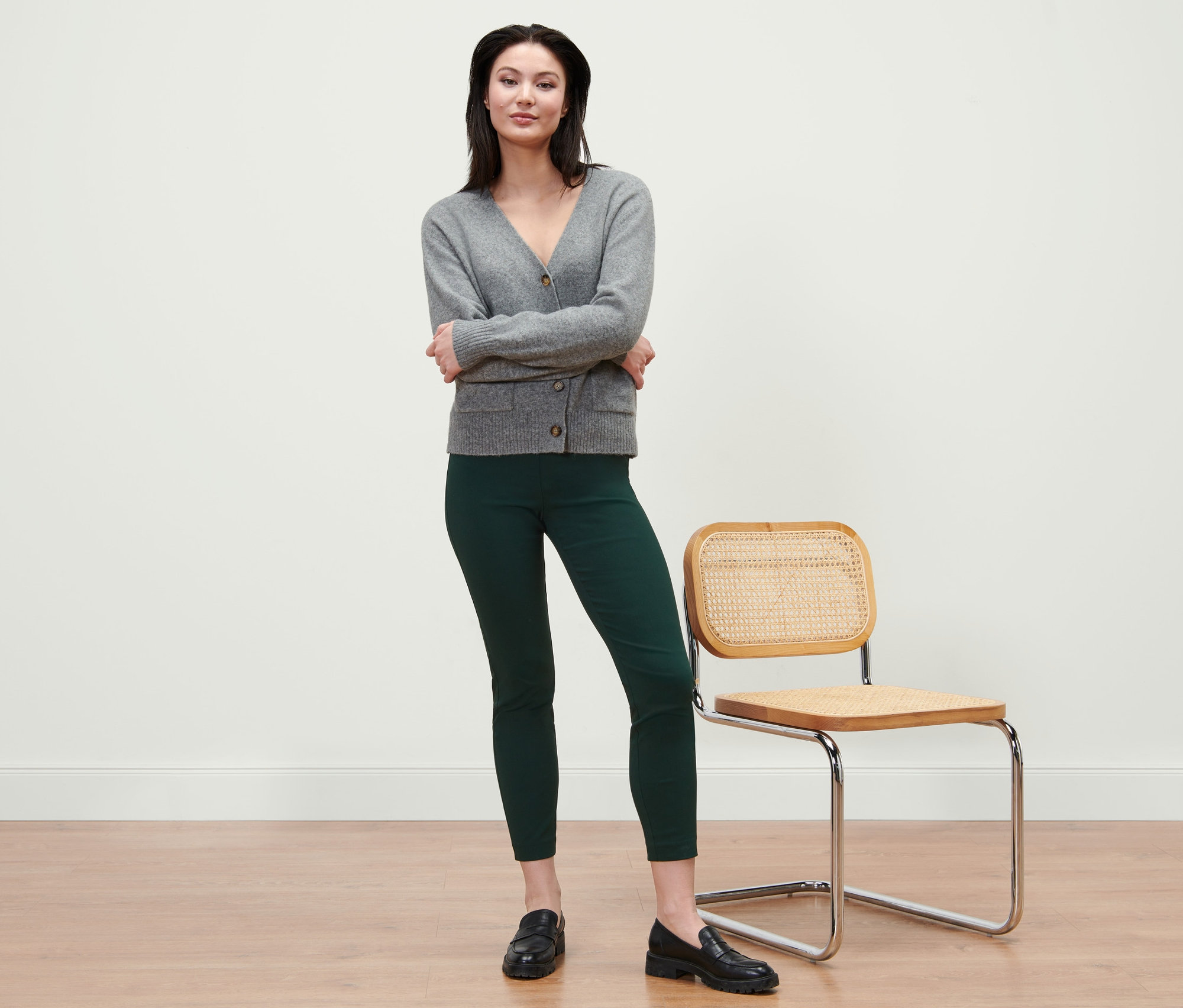 Une femme debout, les bras croisés, portant un cardigan gris et un pantalon stretch vert.
