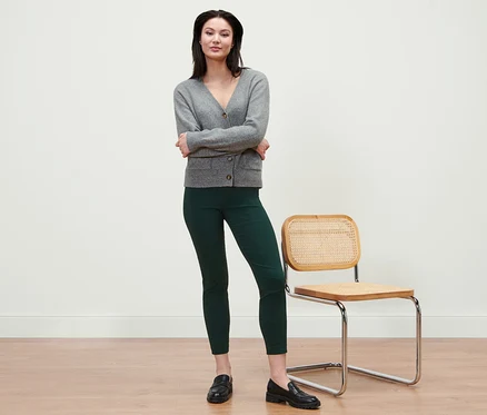 Une femme debout, les bras croisés, portant un cardigan gris et un pantalon stretch vert.