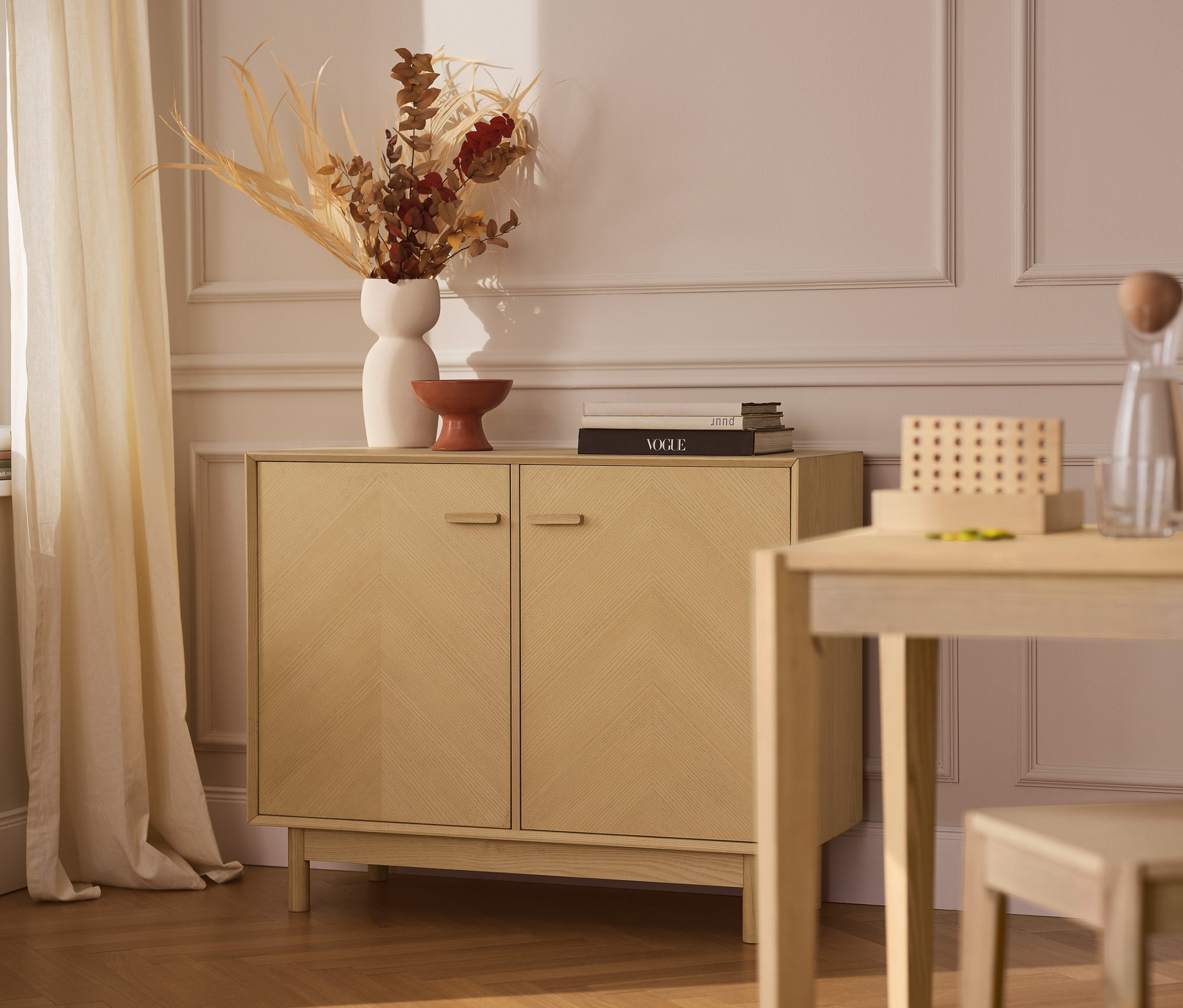 Un buffet en frêne avec des façades de portes décoratives est placé contre un mur. Un vase et des décorations sont posés sur le buffet. Une table et une chaise sont placées à côté du buffet.